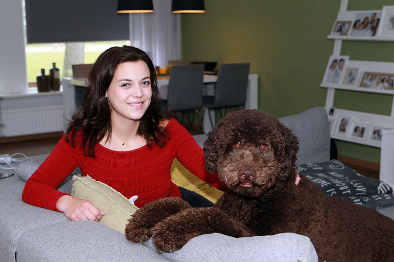 Vrouw met donker haar en rode trui zit lachend op grijze bank naast grote krullende hond. Achtergrond: eettafel, stoelen, rek.