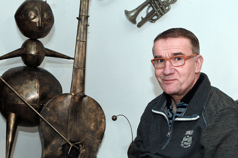 Man met kort haar en bril zit binnen naast metalen beeld van muzikant; trompet hangt aan de muur.