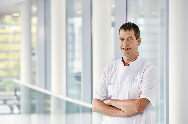 Man in witte doktersjas staat binnen met armen over elkaar en glimlacht naar de camera in een heldere, moderne ruimte.