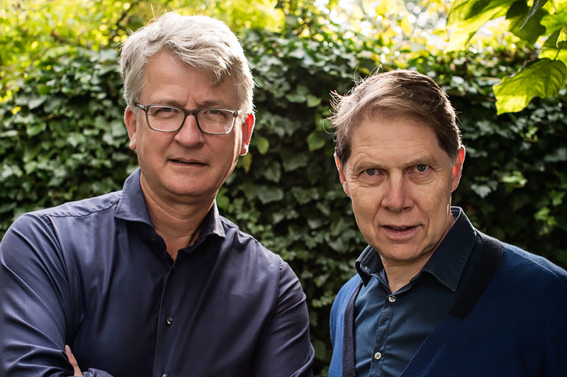 Twee mannen van middelbare leeftijd staan buiten voor een muur met groene klimop. Beiden dragen donkere overhemden.