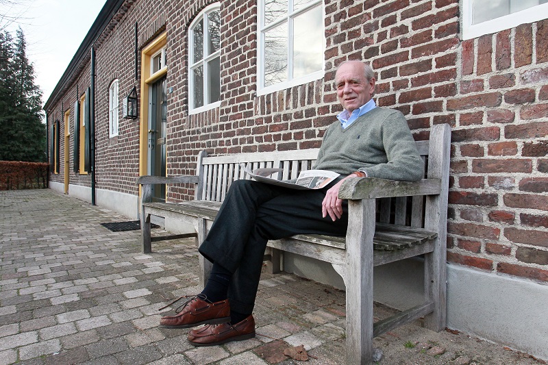 Een oudere man, oud-vaatchirurg, zit op een houten bank buiten bij een bakstenen gebouw met een krant op schoot.