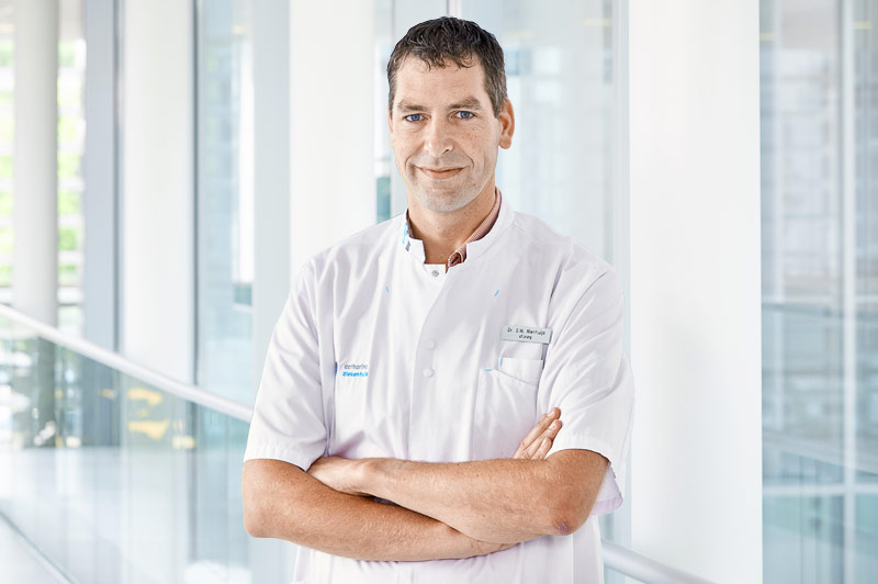 Een man in een witte labjas staat met gekruiste armen in een moderne, lichte gang met grote ramen op de achtergrond.