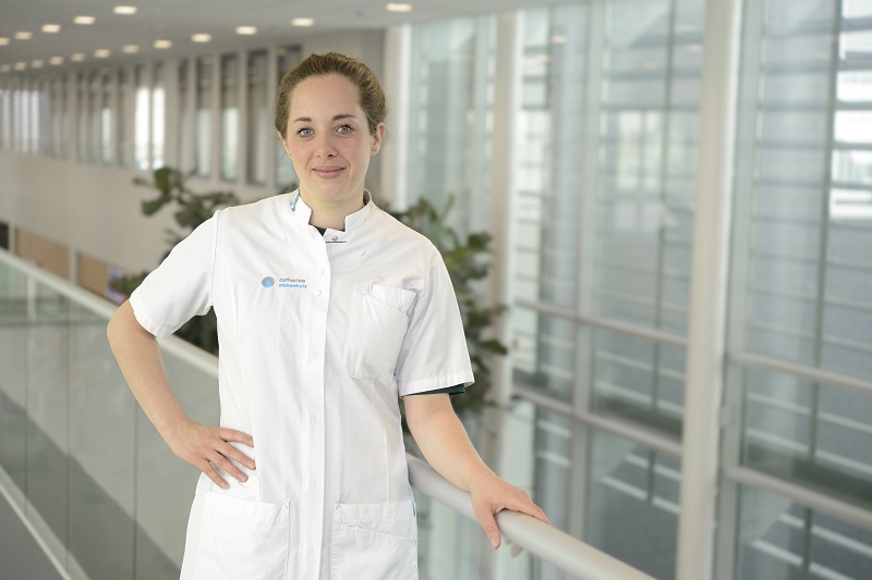 Vrouw in witte medische kleding staat bij een railing in een modern gebouw met grote ramen en planten.