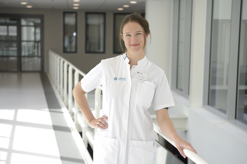 Een promovendus in witte doktersjas staat binnen bij een reling in een lichte bende en kijkt glimlachend naar de camera.