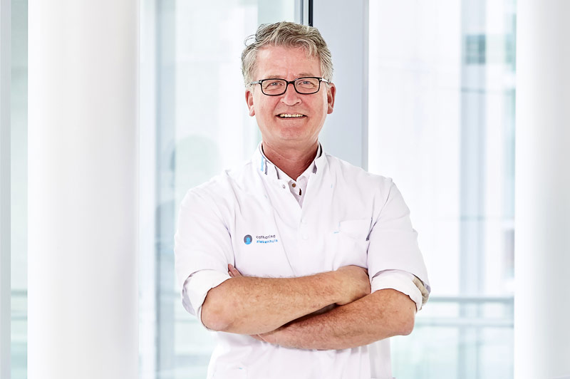 Een man met bril en witte labjas staat binnen met gekruiste armen en glimlacht. De heldere achtergrond heeft grote ramen.