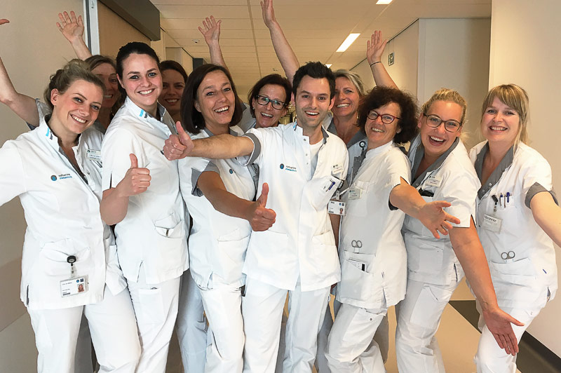Een groep zorgmedewerkers in witte uniformen staat lachend in een ziekenhuisgang en geeft duimen omhoog.