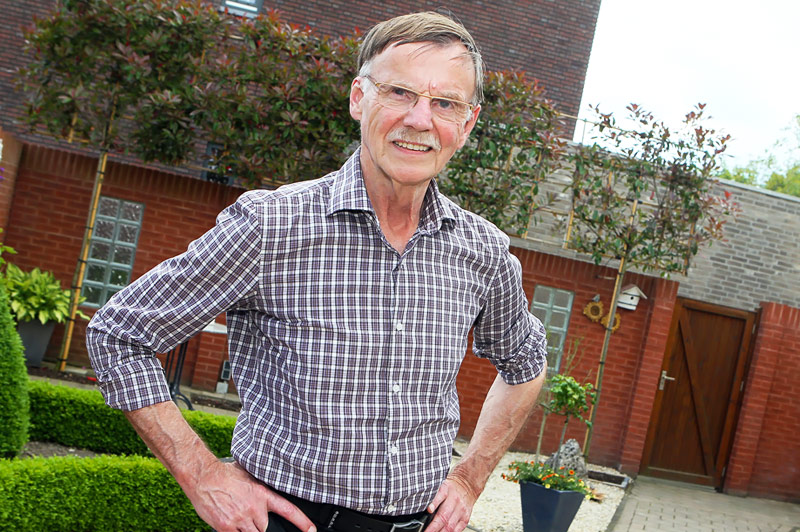 Oudere man met bril en geruite blouse staat in een tuin met zijn handen op zijn heupen, bij een bakstenen gebouw en planten.