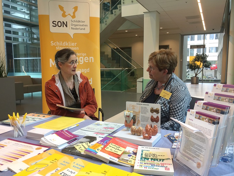 Twee vrouwen bij een infobalie van Schildklier Organisaties Nederland met folders en materialen op tafel over schildklieraandoeningen.