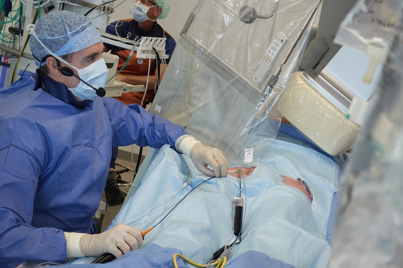 Topcardioloog voert procedure uit op patiënt, draagt beschermende kleding en spreekt via headset in opleidingscentrum.