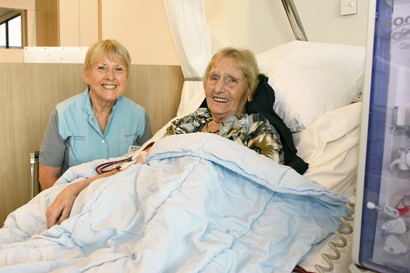 Een oudere vrouw ligt lachend in een ziekenhuisbed naast een zuster; vrijwilligers en medische apparatuur zijn te zien.
