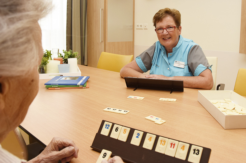 Twee oudere vrouwen spelen een tegelspel aan tafel; een draagt een uniform en glimlacht. Naast hen liggen speltegels en een doos.