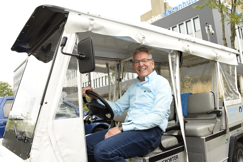 Man met bril en lichtblauw shirt veert 30 jaar vrijwilligerswerk in een elektrische auto voor gebouw met 'Ziekenhuis' op de gevel.
