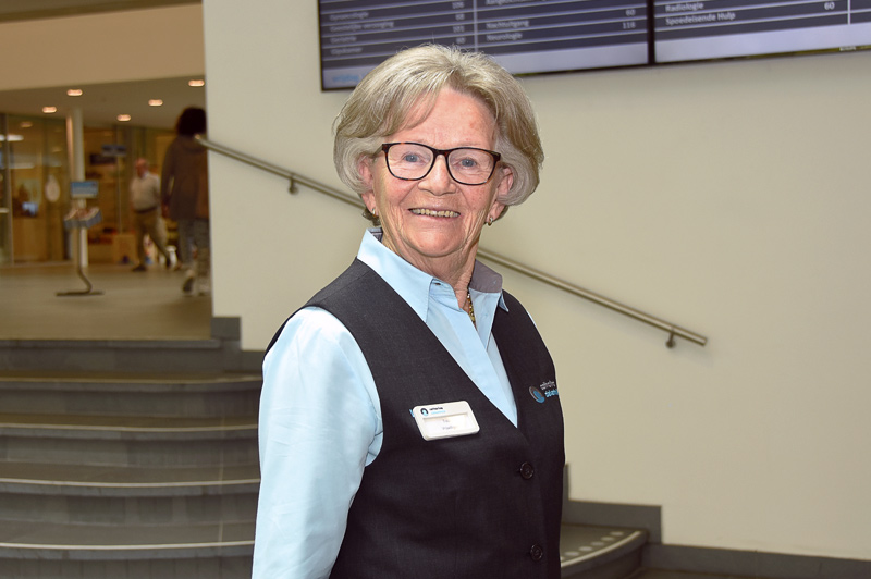 Oudere vrouw met kort grijs haar, bril en naambadge staat binnen bij trap en digitaal bord, veert 30 jaar als vrijwilliger.