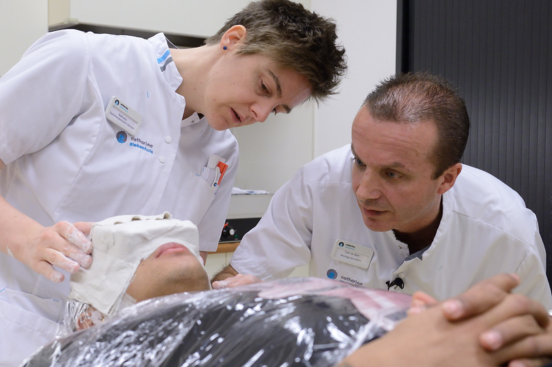 Twee medici, waaronder moulage technicus Felix de Beer, onderzoeken een liggende patiënt met doek op het gezicht en folie op het bovenlichaam.