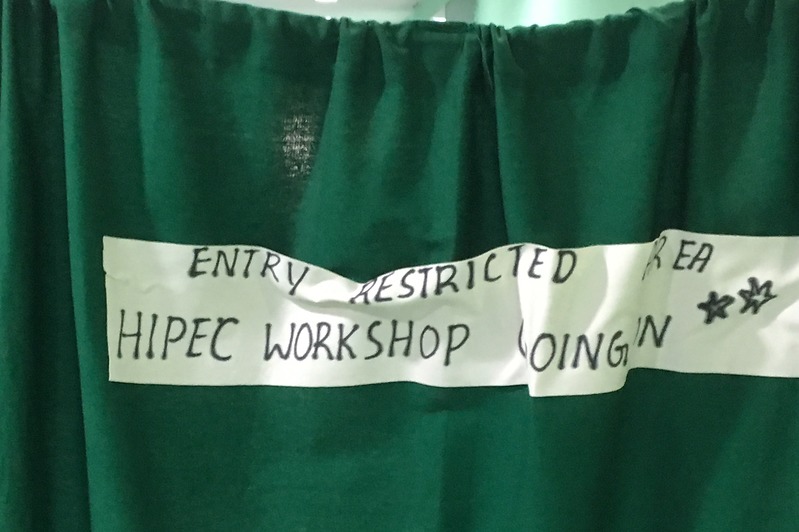 Groen gordijn met handgeschreven bord: ENTRY RESTRICTED AREA HIPEC WORKSHOP GOING IN, toegang beperkt.