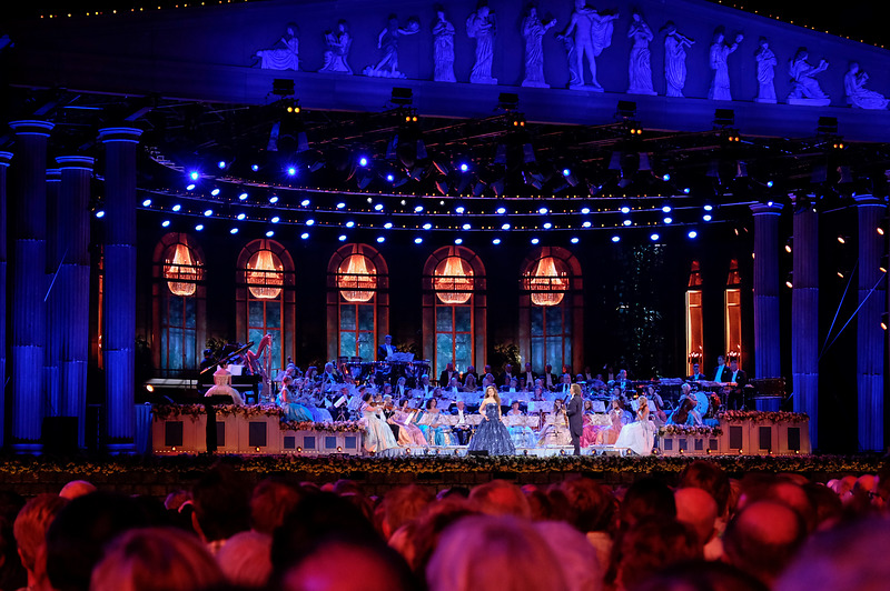 André Rieu dirigeert een symfonieorkest op een verlicht buitentoneel in Maastricht met zuilen en beelden.