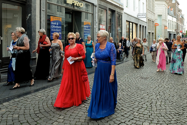 Oudere volwassenen in gala lopen over een Maastrichtse straat, sommigen met papieren, langs winkels tijdens een optocht.