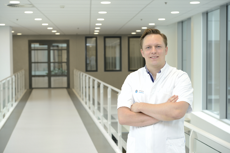 Persoon in witte doktersjas staat met gekruiste armen in een lichte gang, klaar om innovatieve, veiligere operatiemethode te bespreken.