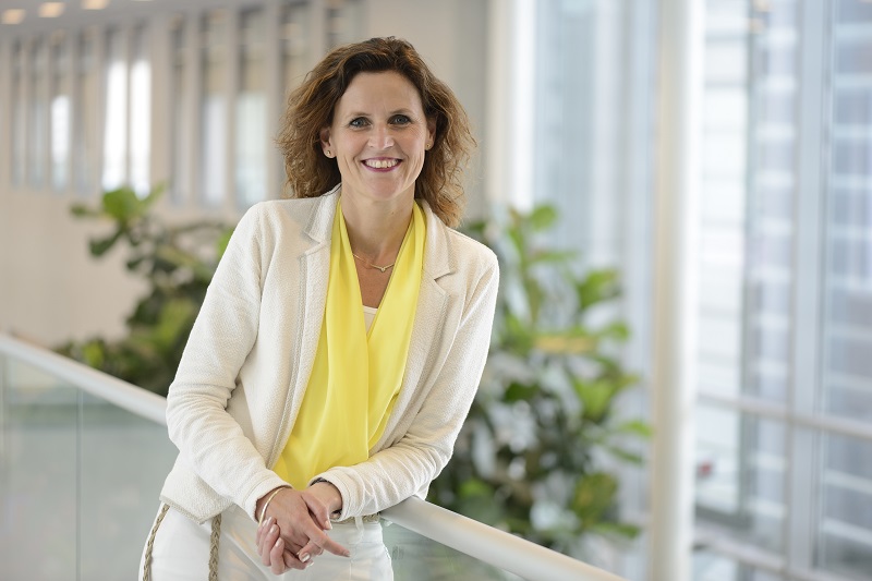 Vrouw met gouden bruin haar in witte blazer en gele blouse, leunend op glazen reling, met planten en grote ramen achter haar.