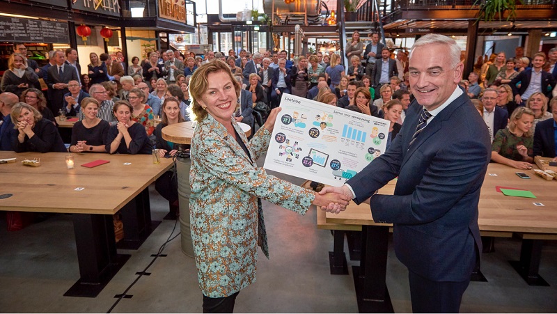 Een man en vrouw in zakelijke kleding schudden handen en tonen een presentatiebord voor een groot zittend publiek binnen.