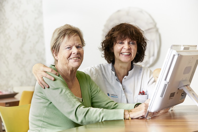 Twee vrouwen aan een tafel, één in groene trui, één in witte zorgkleding, bij een computer in het Catharina Ziekenhuis Nederland.