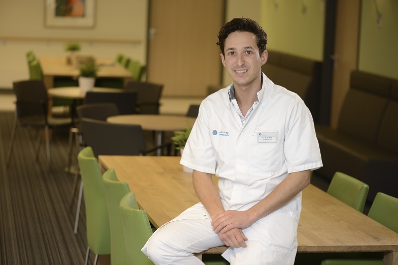 Man in witte medische kleding zit op tafel in felverlichte kamer bij Catharina Ziekenhuis, lachend. Groene stoelen, planten.
