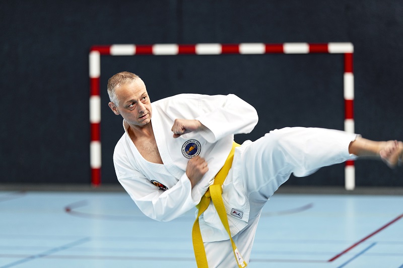 Man in wit vechtsportpak met XXL-band doet zijwaartse trap in gymzaal, rode en witte doelpaal op de achtergrond.