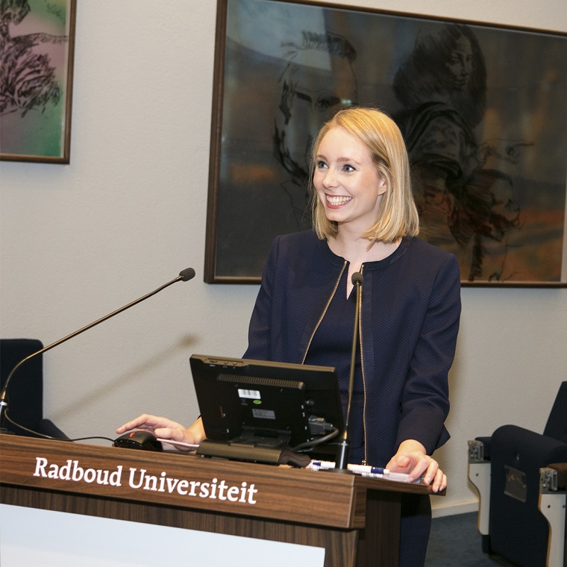 Vrouw in nette kleding spreekt glimlachend bij Radboud Universiteit-podium over vroegtijdige detectie van HPV-tumoren.