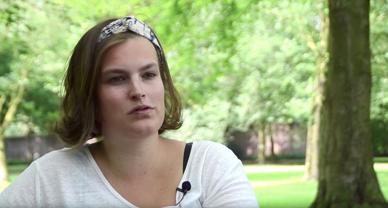 Vrouw met bruin haar, witte top en hoofdband zit buiten bij bomen en gras. Microfoon op shirt; ze vertelt over maagverkleining.