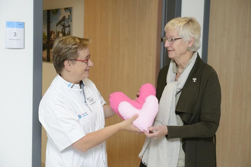 Zorgverlener in witte jas geeft roze hartkussen aan oudere vrouw met bril en sjaal in een bende, als steun voor borstkankerpatiënten.