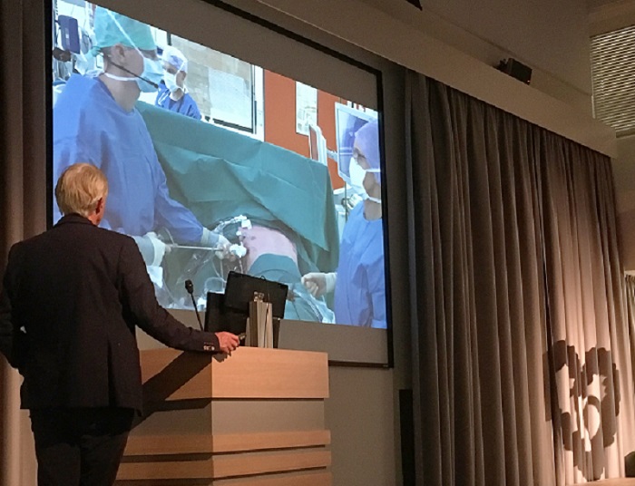 Persoon in pak bij een spreekgestoelte kijkt naar livebeelden van chirurgen in operatiekamer Catharina ziekenhuis tijdens conferentie.