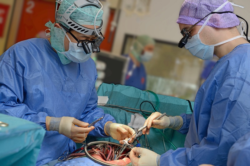 Twee chirurgen in blauwe kleding voeren een openhartoperatie uit, met assistentie van medisch personeel op de achtergrond.