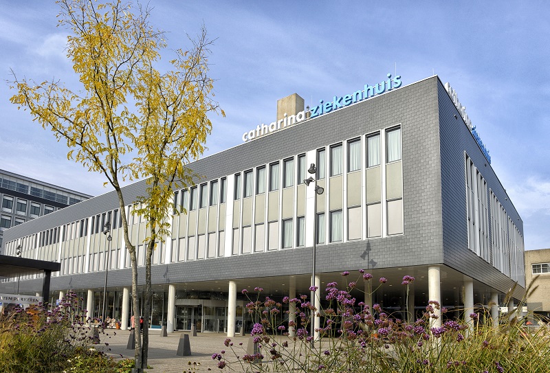 Grijs gebouw met grote ramen en Catharina Ziekenhuis op het dak. Voorgrond: bloeiende planten, boom, blauwe lucht.