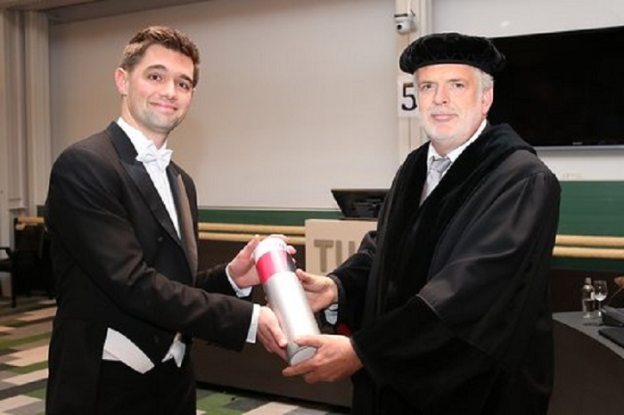 Twee mannen in academische kleding binnen; een geeft een cilindrisch object aan de ander. Ze kijken lachend naar de camera.
