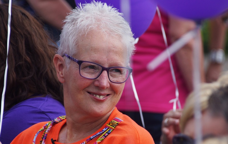 Oudere persoon met kort wit haar en paarse bril lacht; draagt oranje shirt en kleurrijke lanyard. Paarse ballonnen op achtergrond.