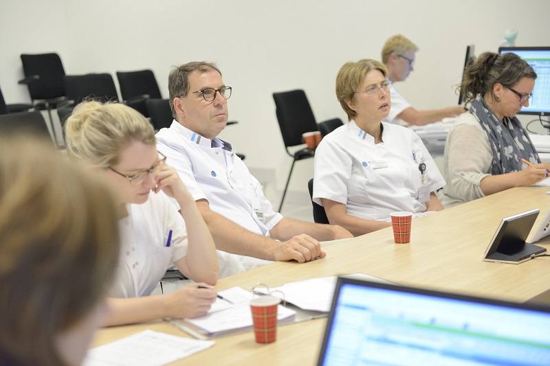 Vier zorgverleners, drie in witte uniformen en één met grijze sjaal, overleggen aan tafel over gynaecologische oncologie.