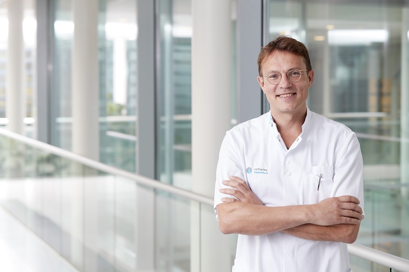 Arts met bril en witte jas staat met gekruiste armen in lichte, moderne hal, klaar om preventie en behandelingen te bespreken.