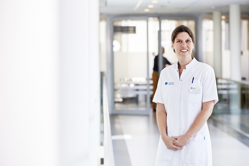 Zorgprofessional in wit uniform in lichte hal van Catharina Ziekenhuis, met glazen wanden en vage figuren op de achtergrond.