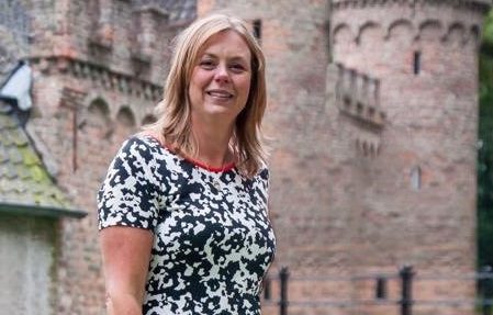 Vrouw met blonde haar en zwart-witte jurk staat buiten voor bakstenen gebouw met bogen, klaar voor een huwelijksspeech.