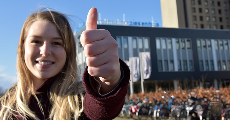 Jonge vrouw voor modern gebouw geeft duim omhoog tijdens een evaluatie in 2018. Op de achtergrond fietsen en vlaggen.