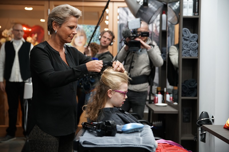 Een kapper stylet het haar van een meisje met bril in een salon; omstanders en een cameraman kijken toe.