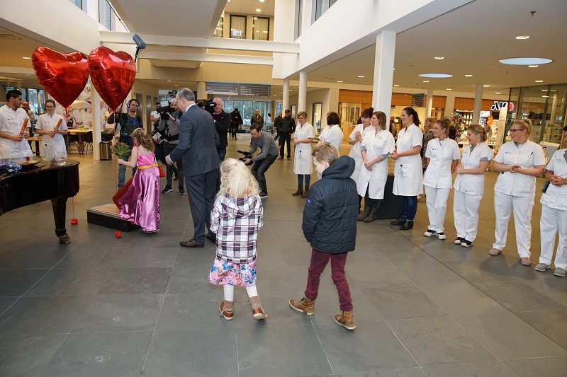 Groep kinderen en volwassenen in witte uniformen in ruime hal. Twee rode hartballonnen en een zwarte piano zijn zichtbaar.