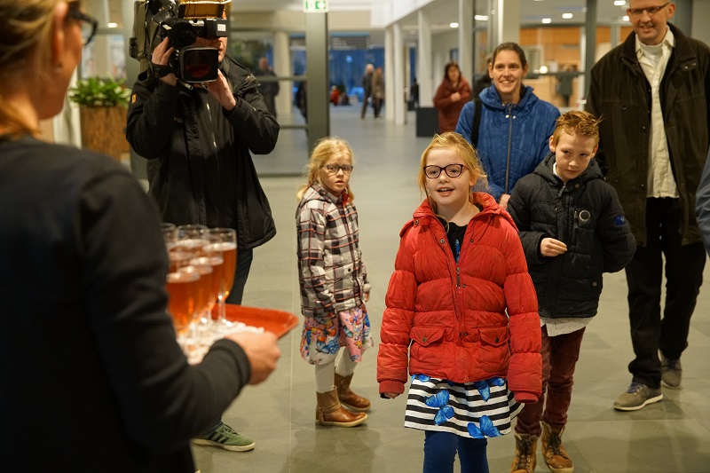 Kinderen en volwassenen staan binnen tegenover iemand met een dienblad drankjes. Een cameraman filmt de groep.