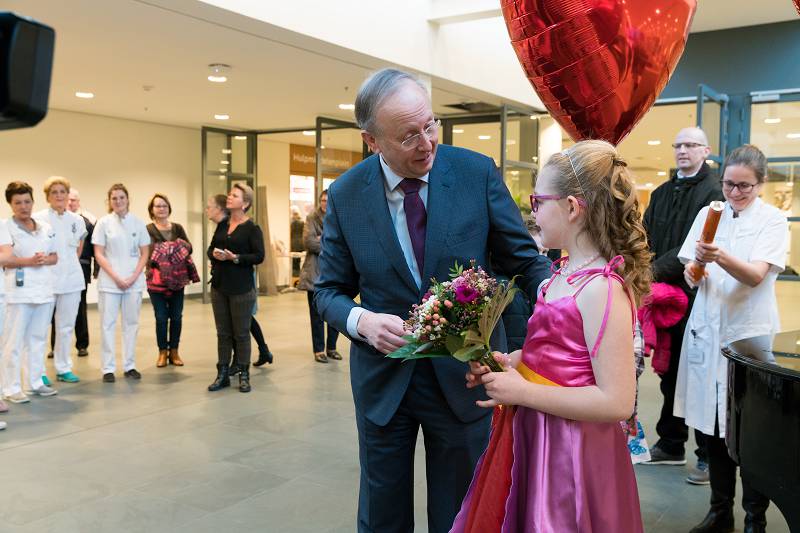 Man in pak spreekt meisje in roze jurk met bloemen en rood hartballon, medisch personeel kijkt toe, binnen in openbaar gebouw.