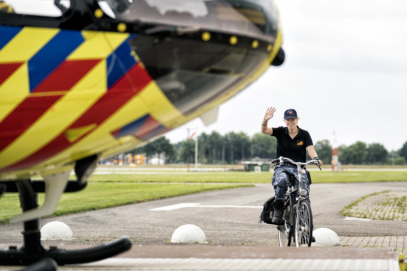 Persoon met donkere trui en huisdier wacht op fiets bij helikopter met rood, geel en blauw, buiten op een bewolkte dag.