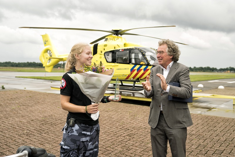 Jonge vrouw in uniform met bloemen wordt toegejuicht door oudere man in pak. Gele traumahelikopter op de achtergrond.
