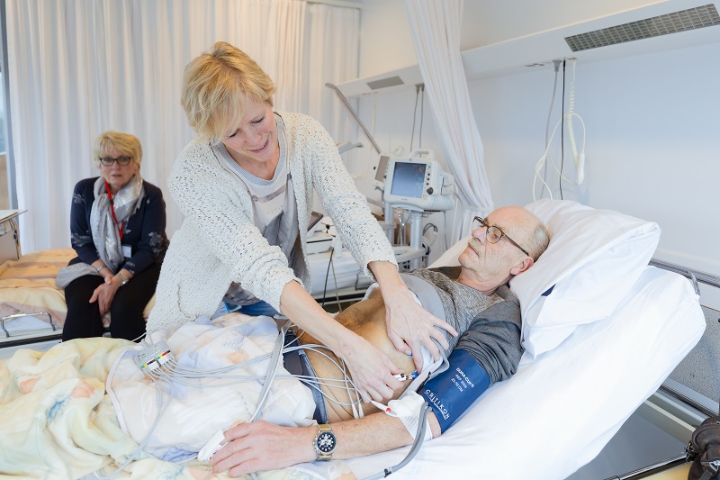 Een chirurg controleert de waarden van een man in bed met monitors; een vrouw kijkt toe. Apparatuur voor beeldgestuurde chirurgie.