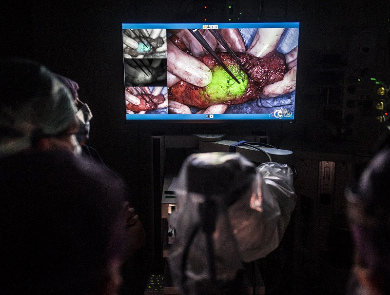 Monitor toont close-up van chirurg bij beeldgeleide chirurgie; groene kankercellen zichtbaar. Medisch team in schemerige kamer.
