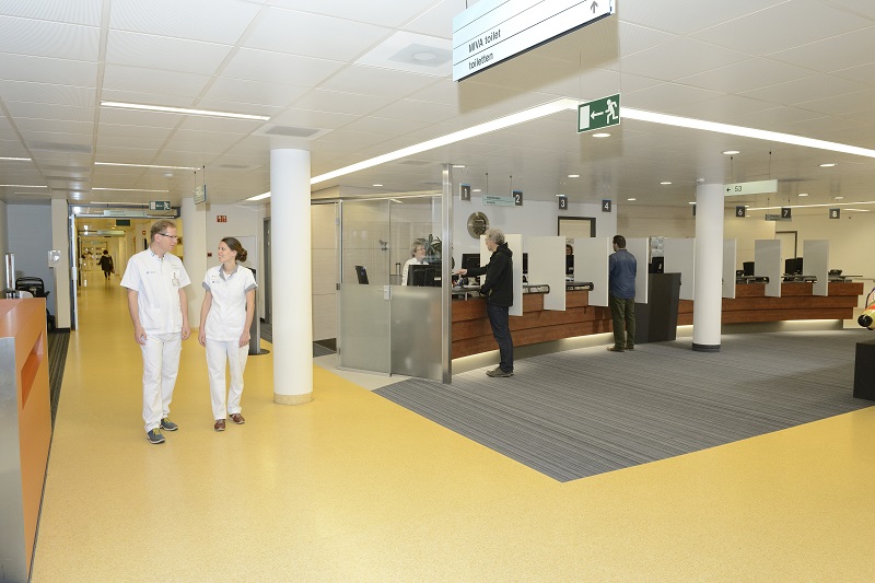 Twee mensen in witte uniformen lopen door een ziekenhuisgang. Op de achtergrond staan mensen bij de receptie.