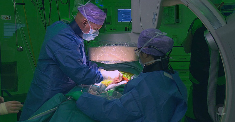 Twee medici in blauwe OK-jassen en paarse mutsen voeren een operatie uit onder groen licht, met medische apparatuur.